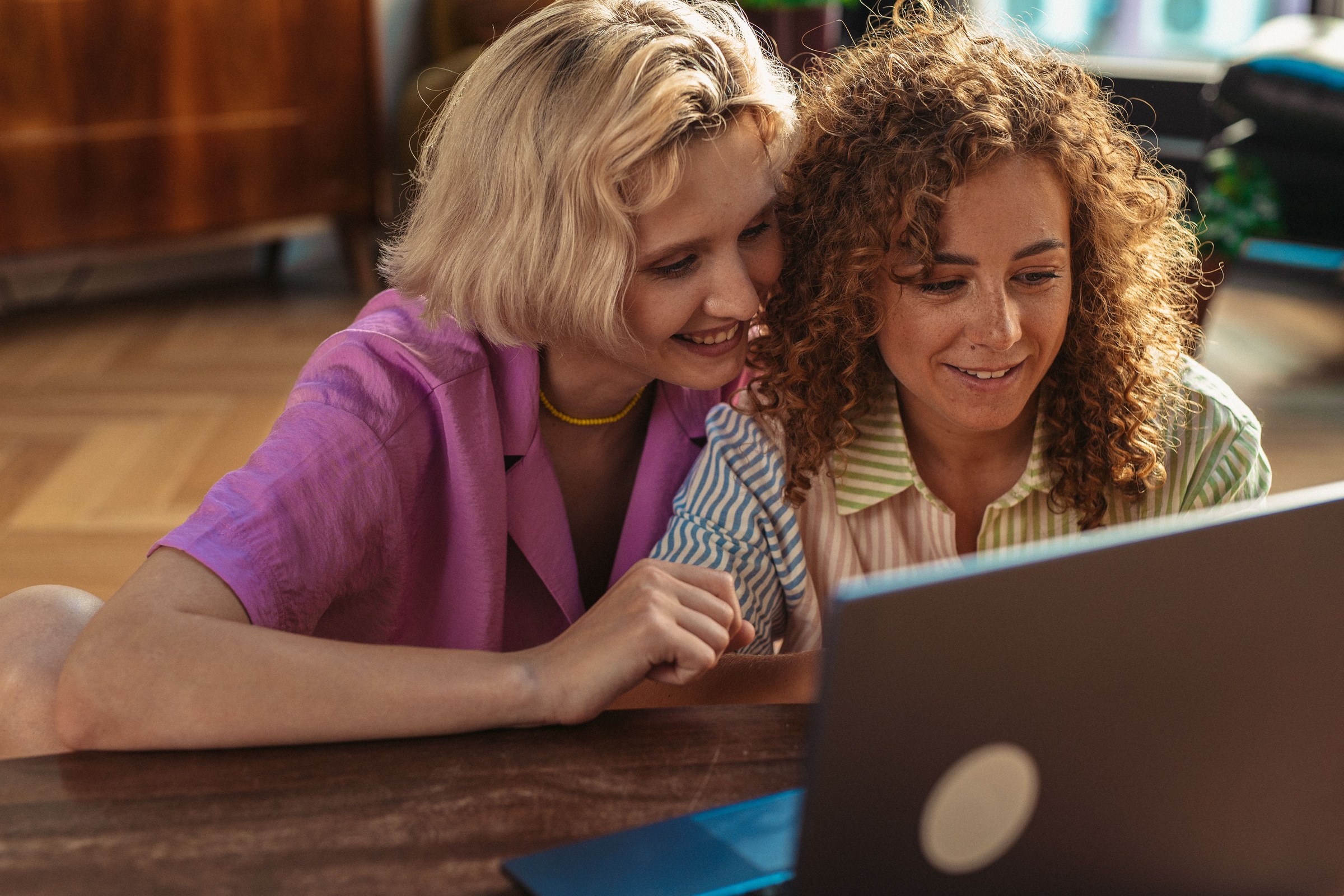 Two smiling, polished businesswomen leaning in together to check which local businesses an AI assistant recommends on a laptop