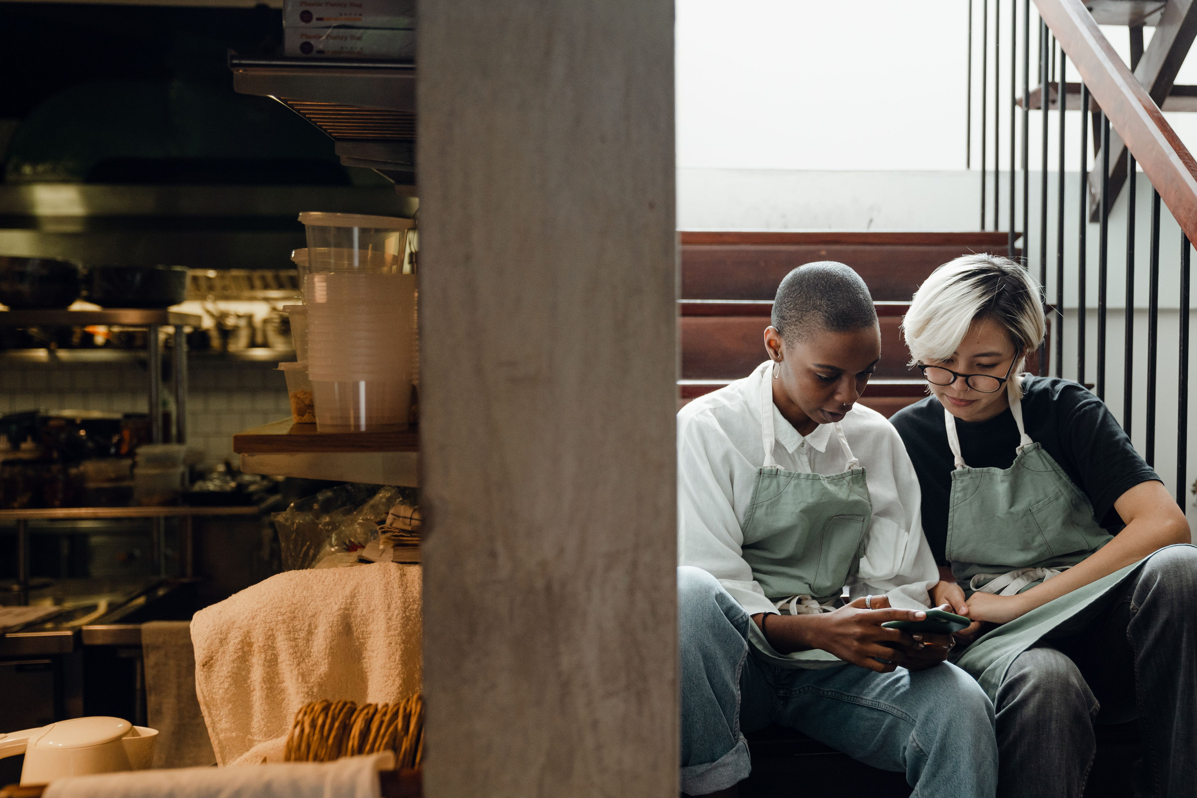 Two cafe workers in green aprons sitting on the back stairs during a break, looking at their Google Business Profile posts on a smartphone