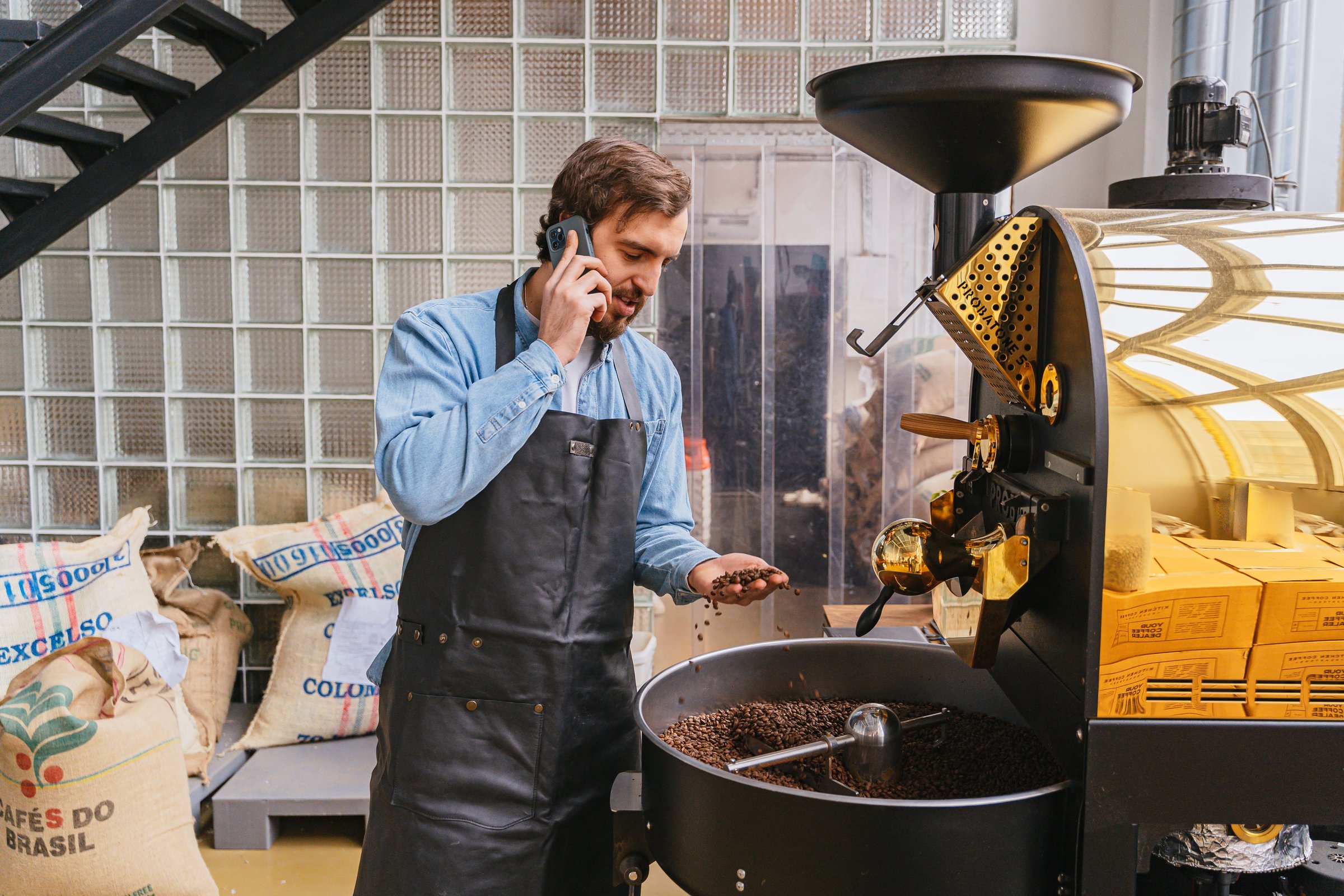 Coffee roaster owner in a work apron on the phone beside his roasting machine, calling a marketing company with his list of questions