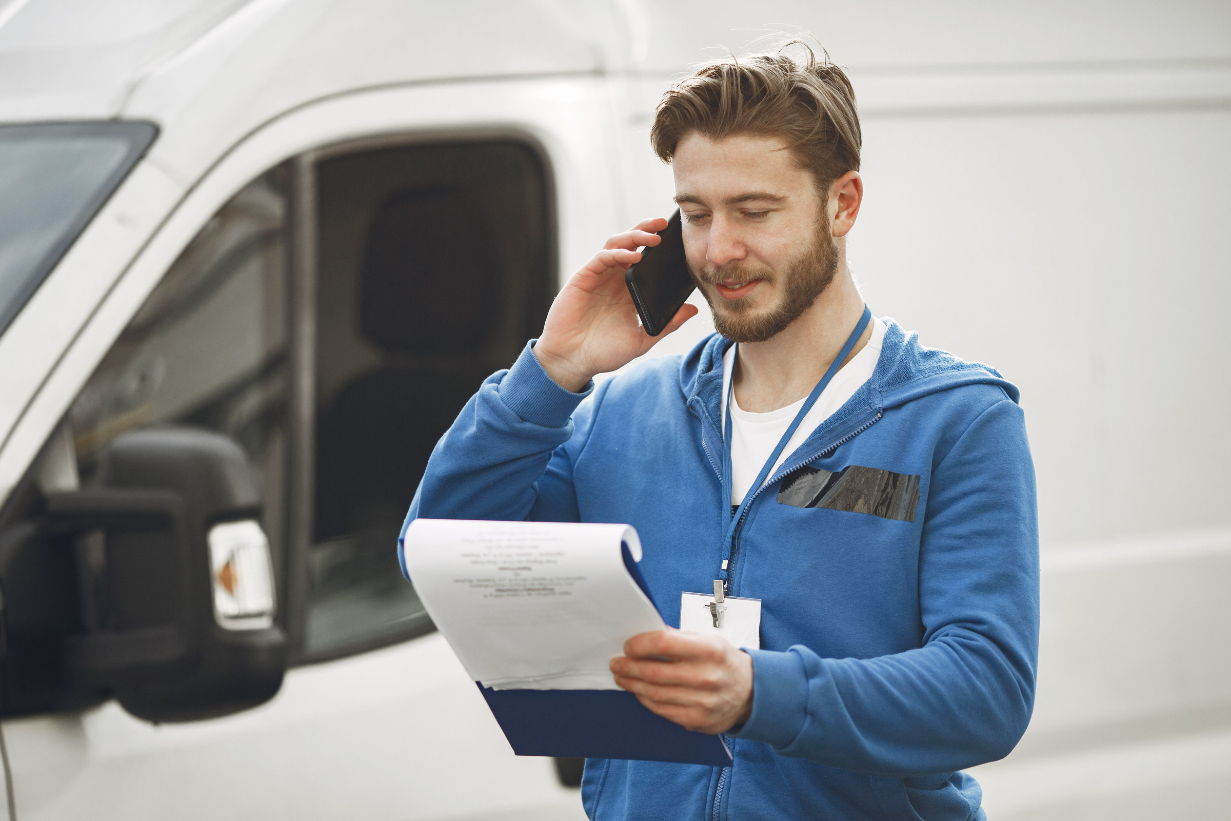 Service worker in a blue hoodie standing beside his white work van, holding a clipboard while calling back a new lead from a Google ad