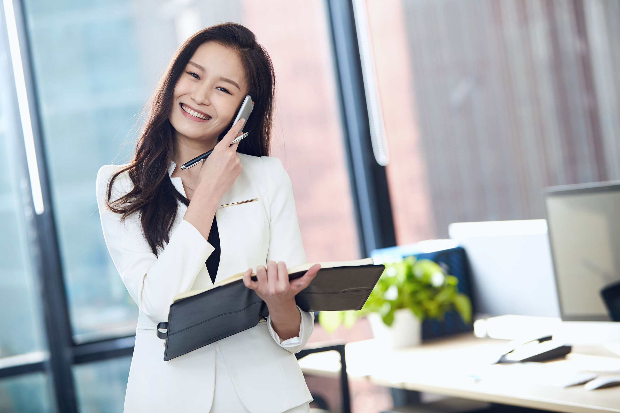 Smiling business owner in a white blazer talking on a smartphone in a bright office, ready to take a call from a camera-search visitor