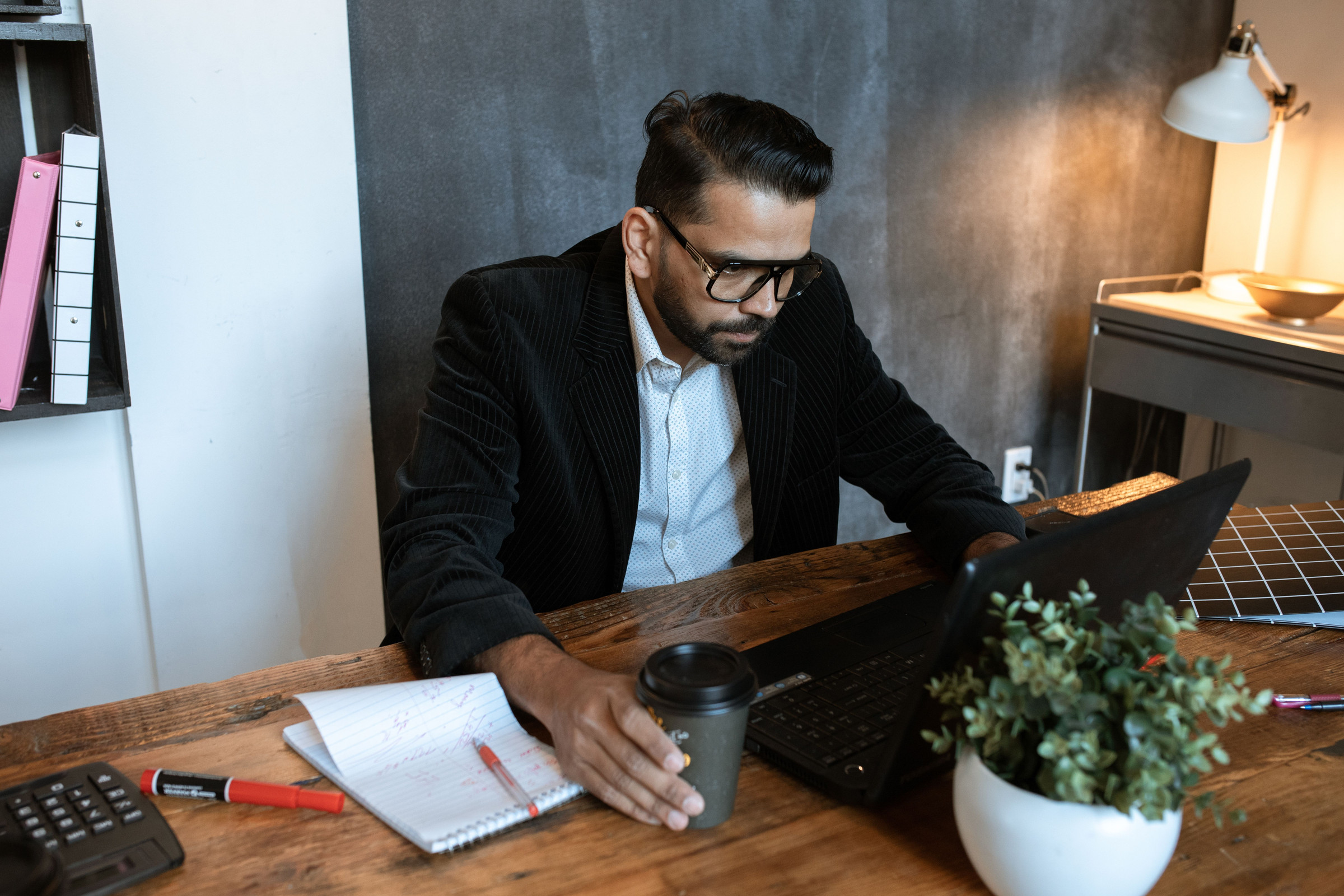 Business owner in glasses and a dark blazer at his office desk, reading a Google ranking report on his laptop with a notebook and coffee beside him