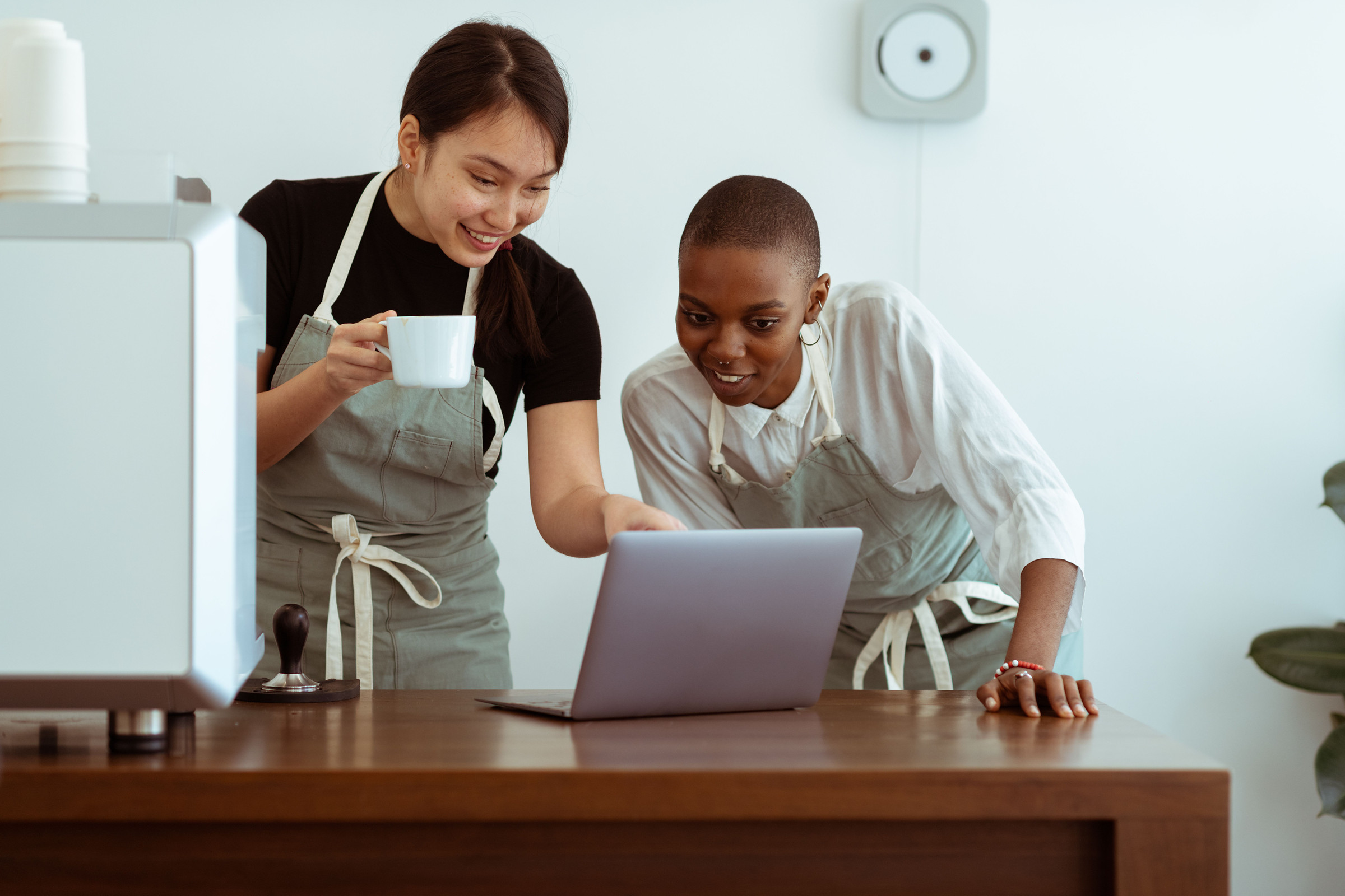 Two smiling, polished café owners in aprons looking at a laptop together at their counter while they update their Google Business Profile