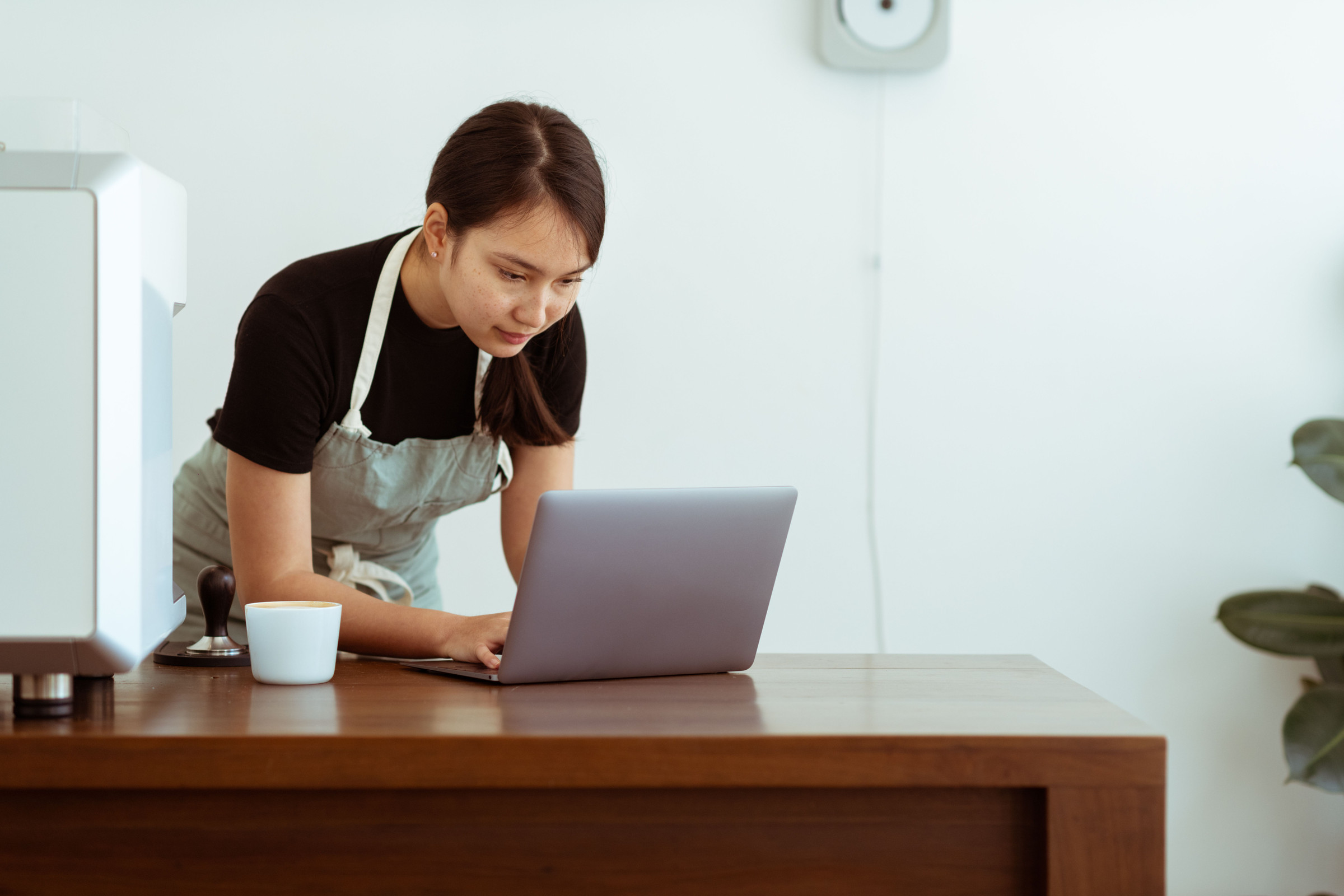 Shop owner in a gray apron leaning over a laptop at her counter, checking her website report for ChatGPT and other AI visits