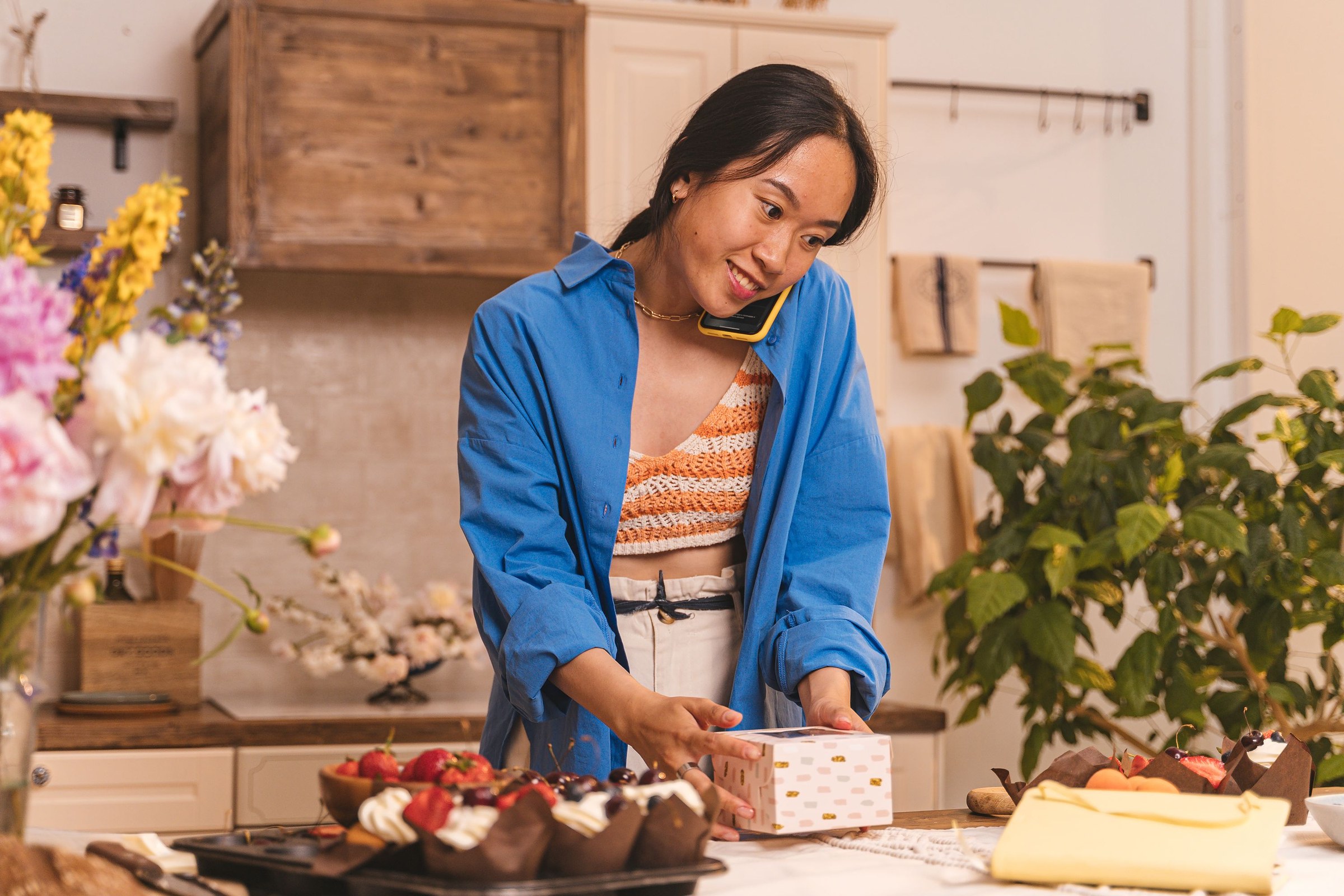 Smiling bakery owner on a phone call while arranging boxed pastries and flowers on the counter, the kind of local business people discover with camera search