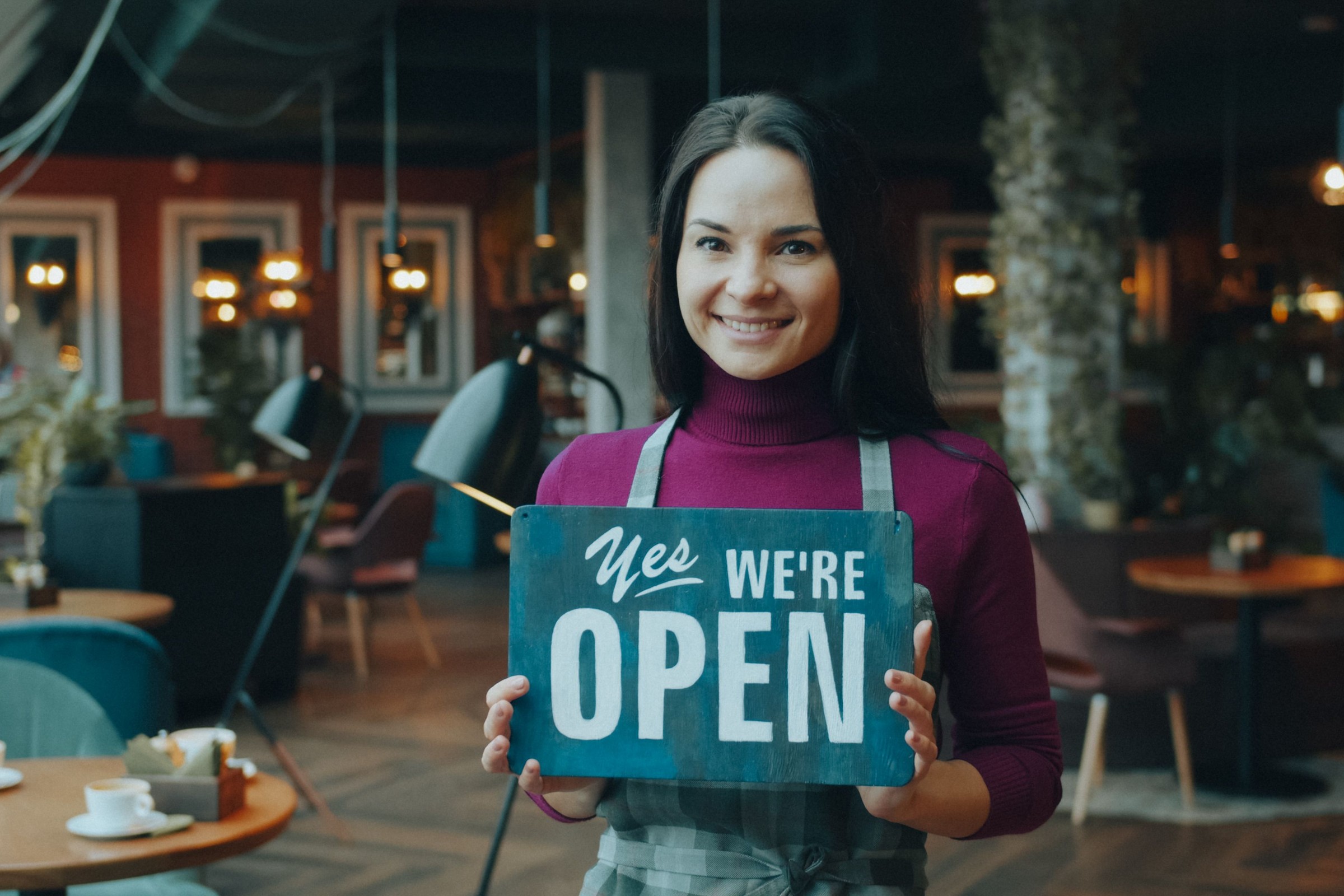Smiling cafe owner holding a Yes We’re Open sign inside the restaurant, representing a local storefront whose Google Knowledge Panel customers check on their phones before they visit