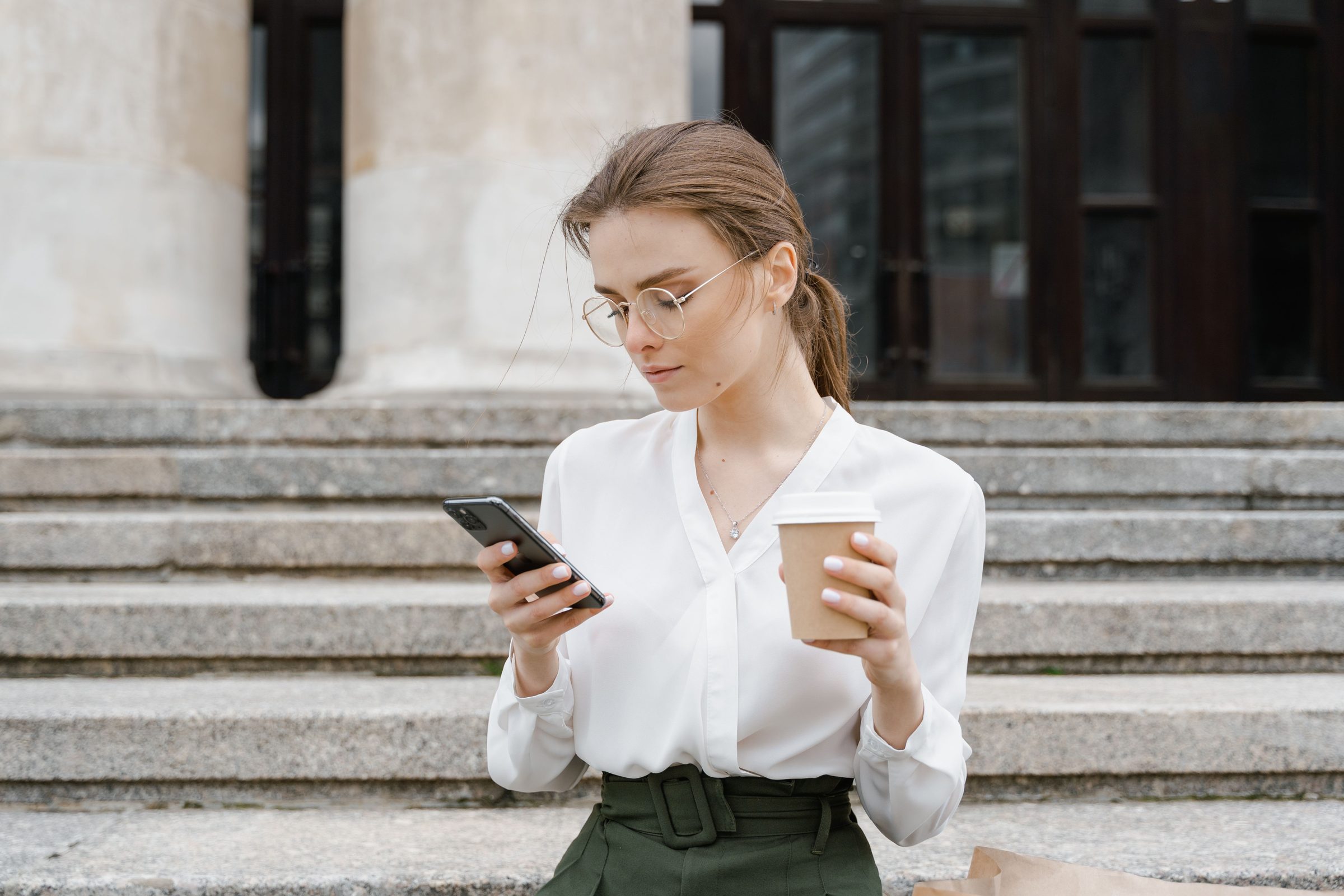 Woman on city steps reading a text message on her phone while holding a coffee