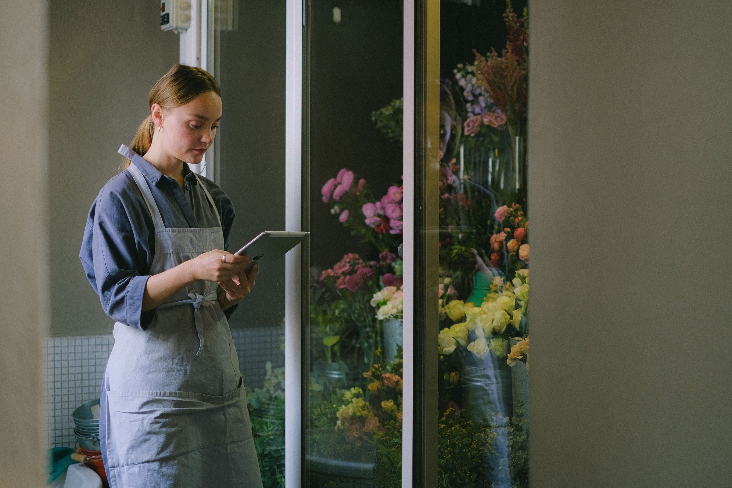 Florist in a blue apron standing at her shop door with a tablet, adding a new post about fresh flowers to her Google Business Profile