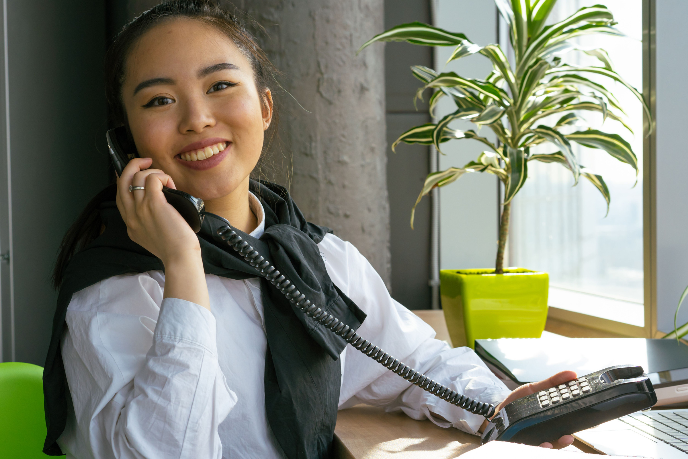 Smiling, polished receptionist answering the front desk phone in a bright office with plants, ready to book the caller