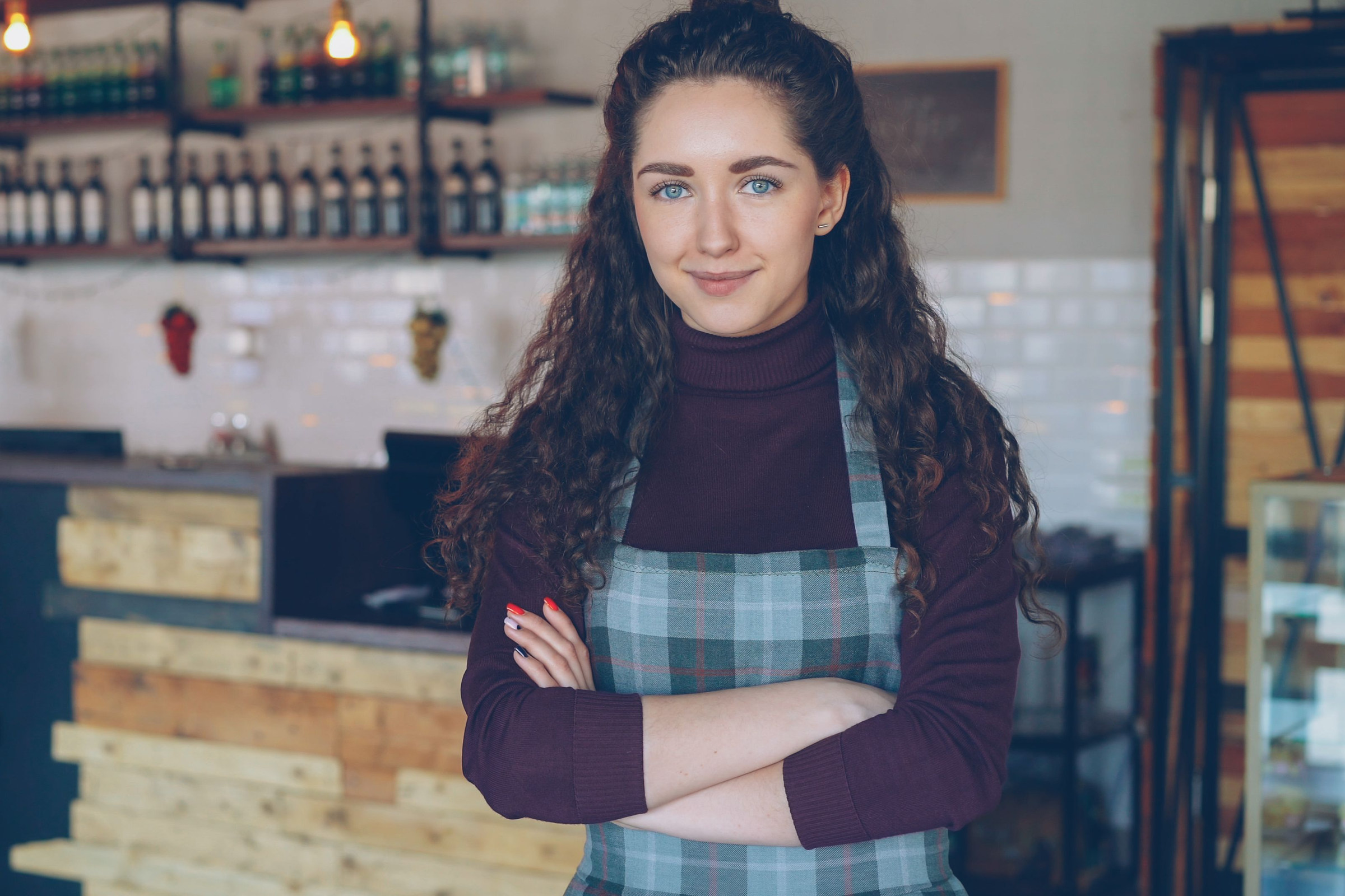 Confident, smiling local café owner in a plaid apron standing with arms crossed in her polished shop, representing a business that earns repeat AI recommendations