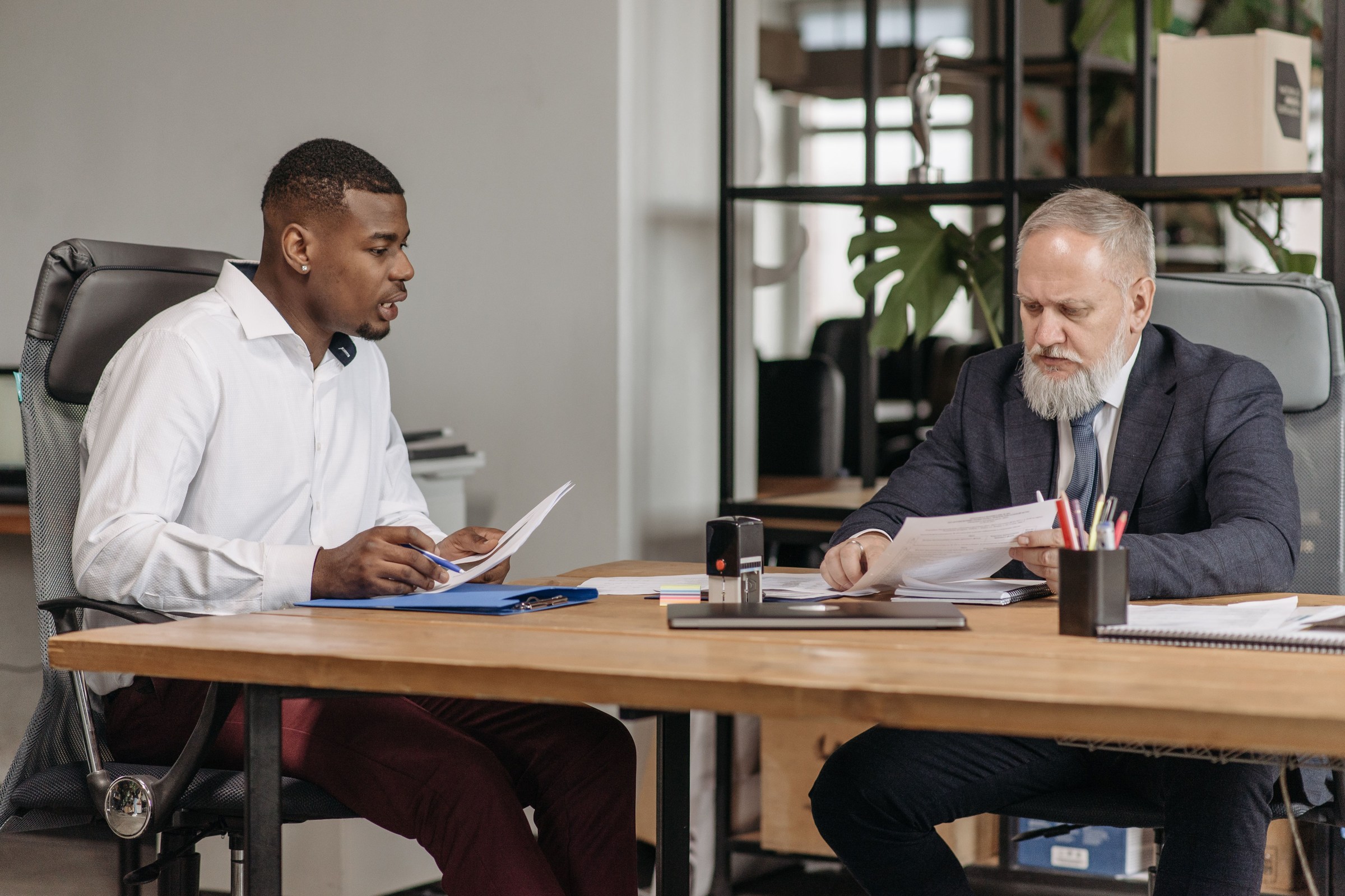 Business owner in a white shirt reading the terms of a marketing contract at a desk while an older advisor in a suit reviews the paperwork across from him