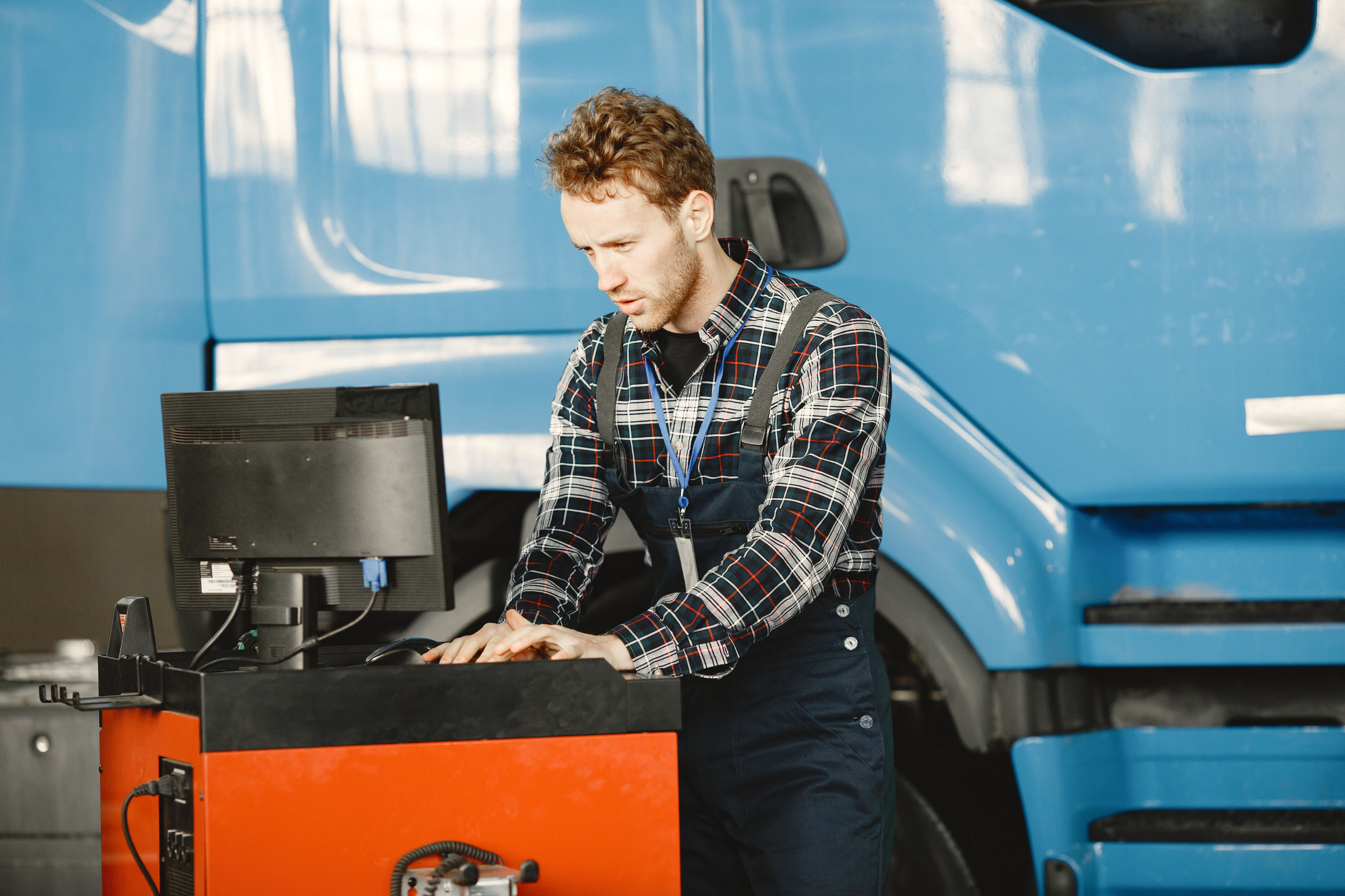Truck repair shop owner in a plaid shirt reading his Google reviews on the shop computer in front of a blue truck