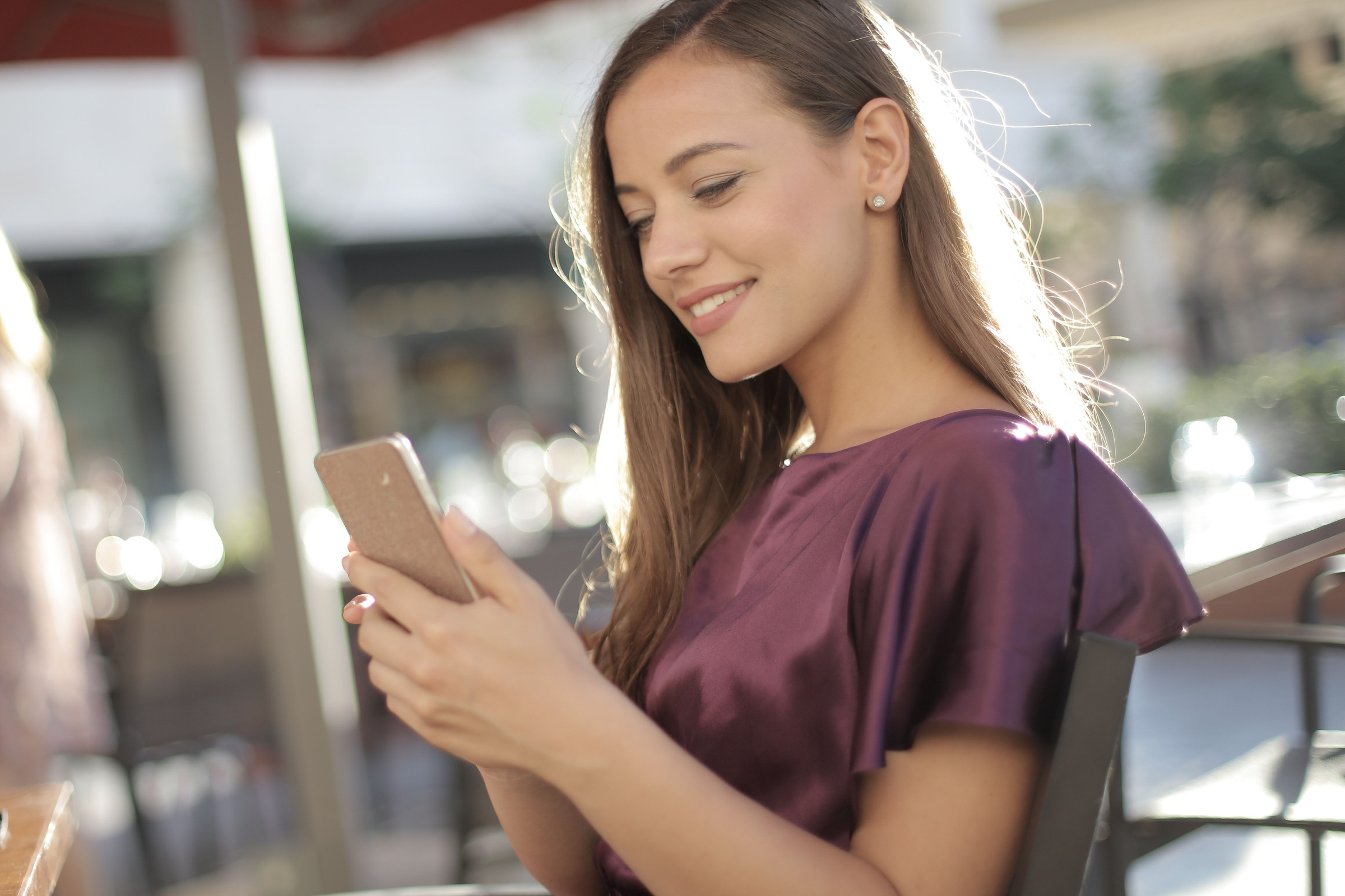 Smiling, polished business owner with long hair texting a customer back on her phone at a sunny café table