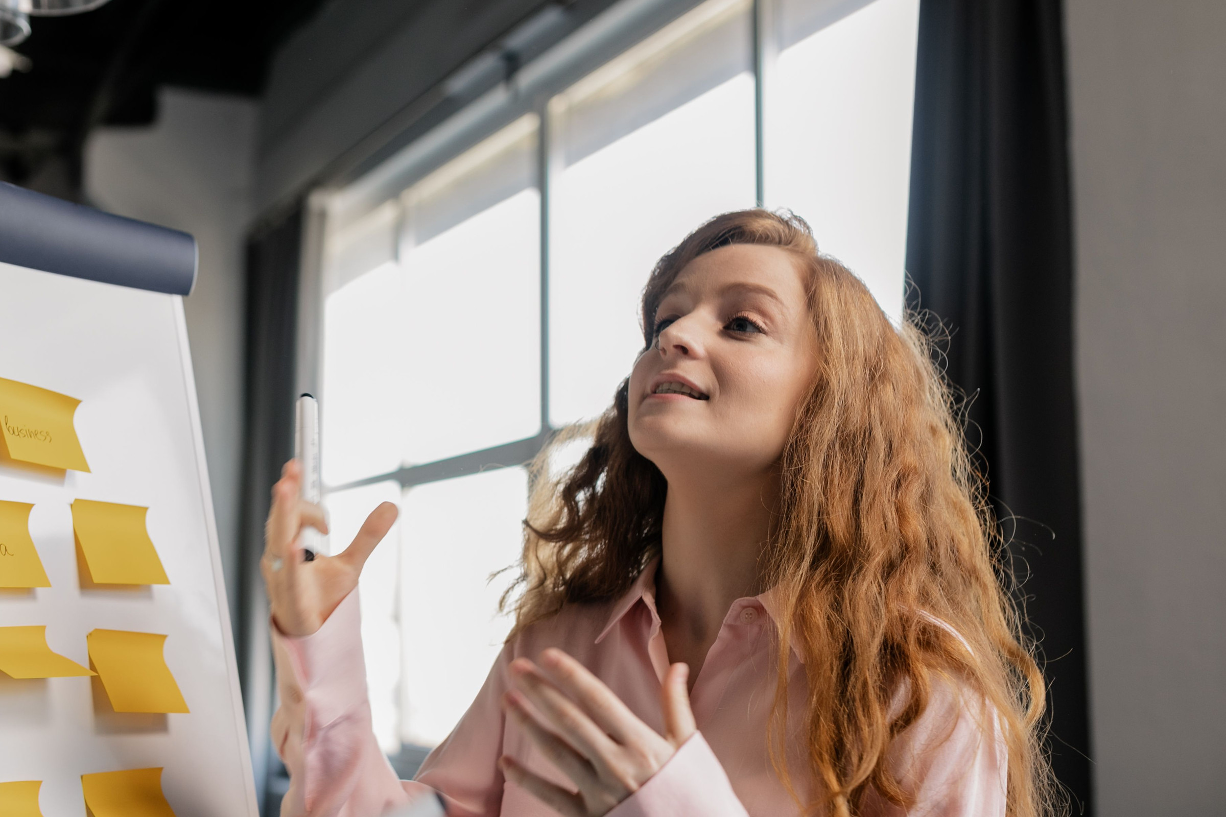 Polished marketing strategist with long red hair explaining an AI visibility plan next to a sticky-note strategy board in a bright office