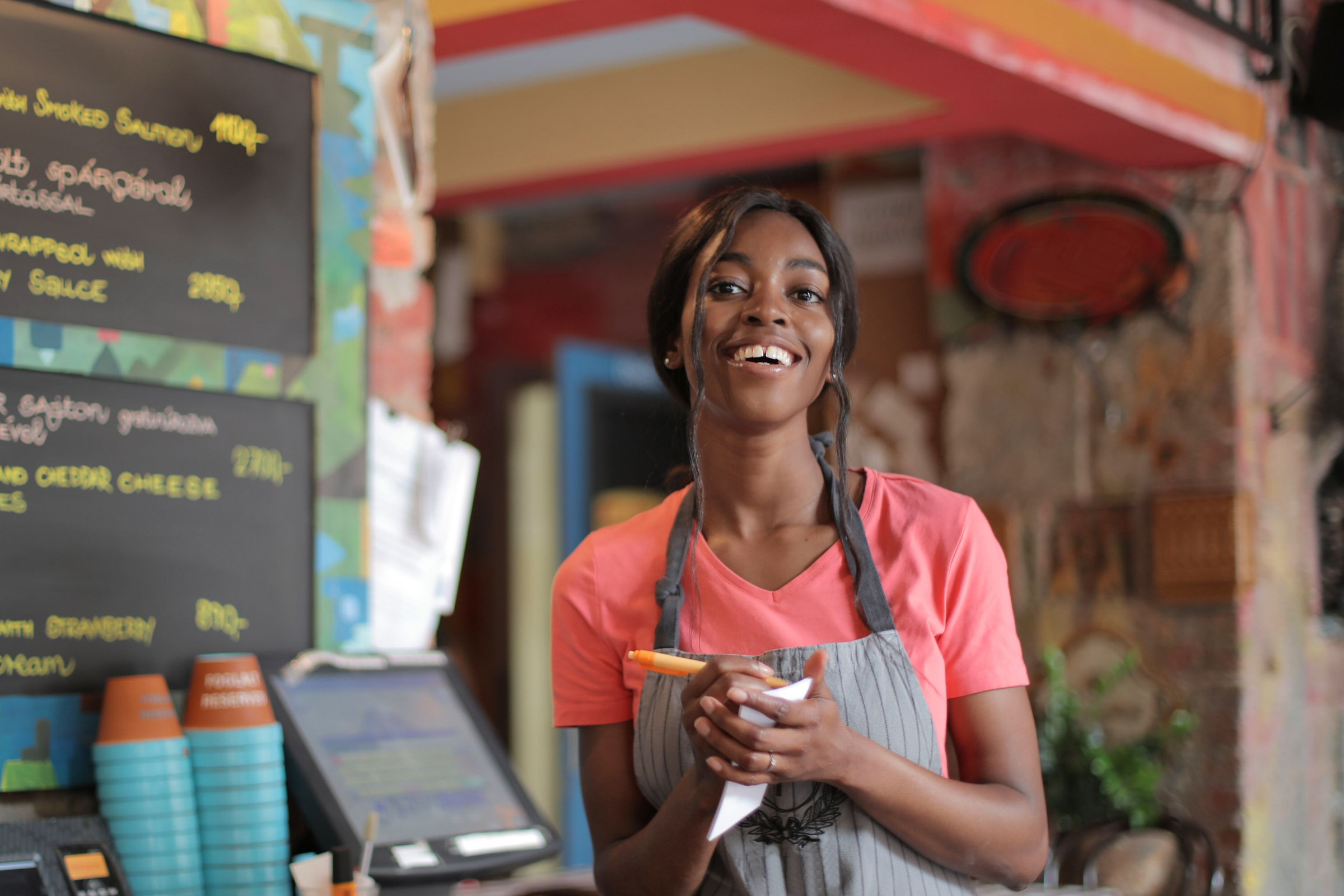 Smiling, friendly café owner in a pink shirt and apron standing by her chalkboard specials menu, the kind of offer that now belongs in a Google post