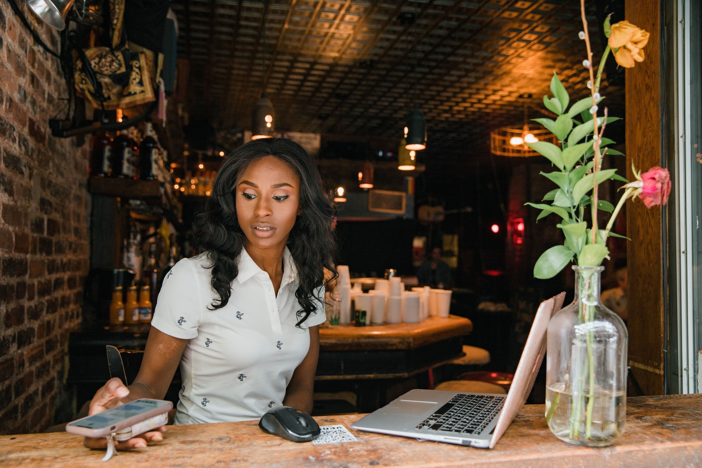 Cafe owner at the counter of her coffee bar checking a new customer review on her phone, with her laptop open beside her