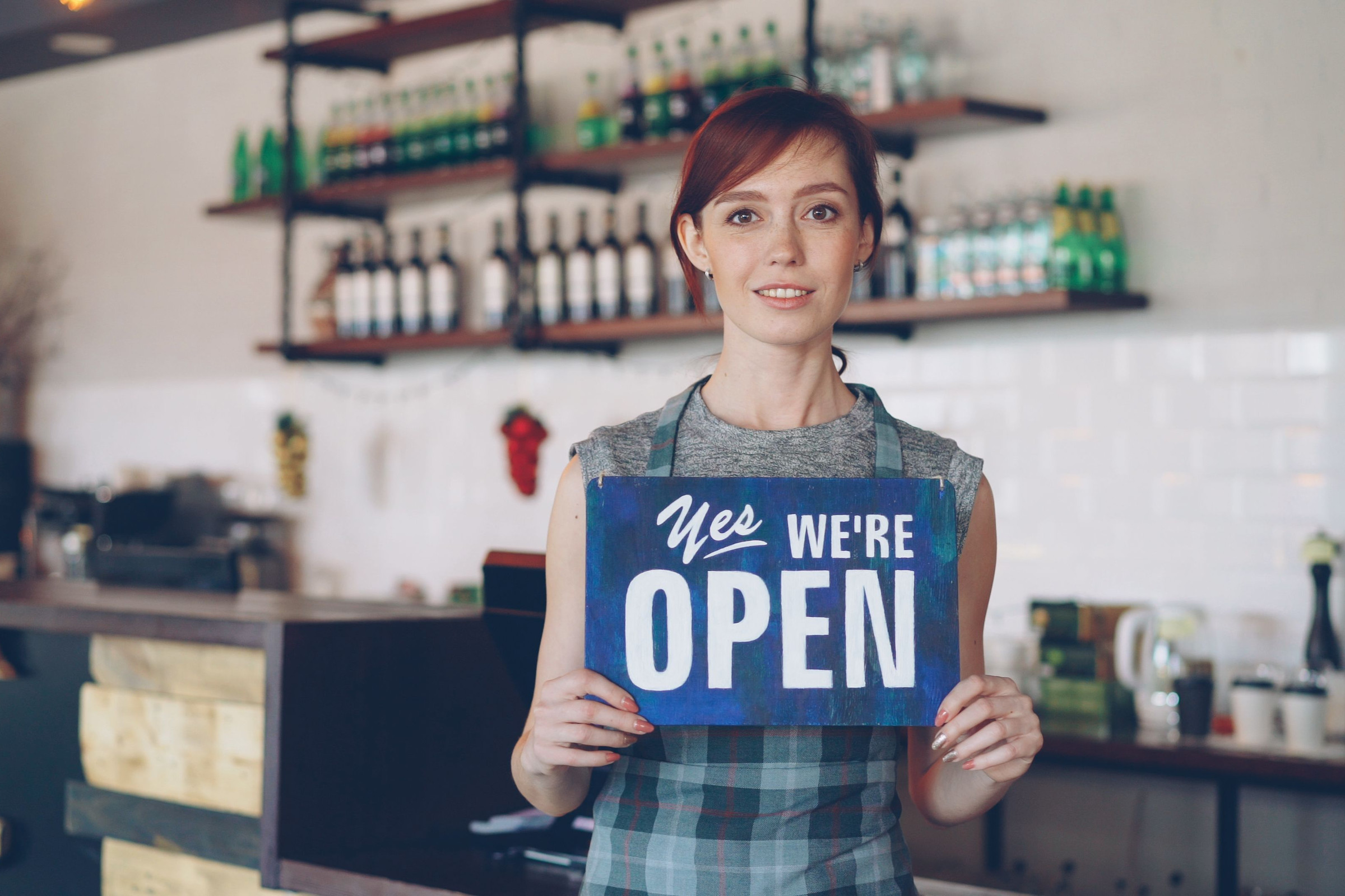 Smiling cafe owner in a plaid apron holding a Yes We Are Open sign inside her shop, the kind of simple real photo that makes a good Google Business Profile post