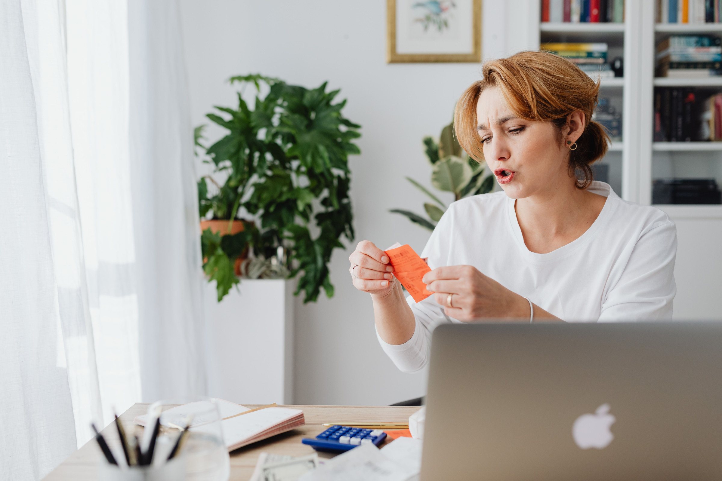 Business owner at a bright home office desk holding a note beside her laptop and calculator, checking how much of her daily ad budget goes to each place her ads show
