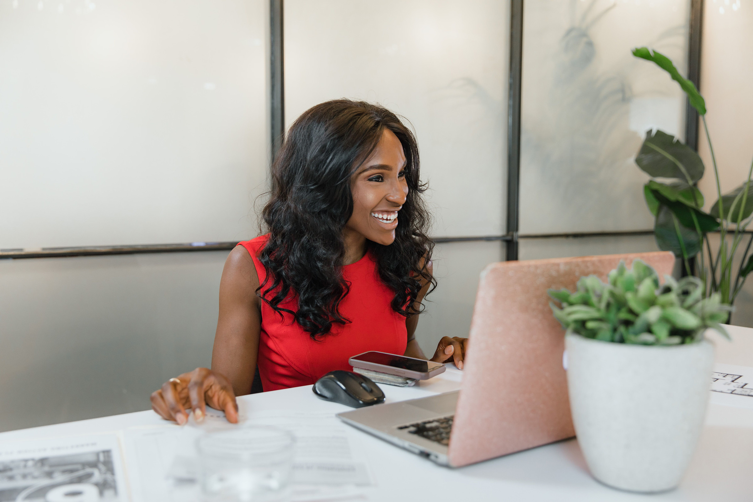 Smiling, polished business owner in a red dress reviewing her weekly lead report on a laptop at a bright white desk