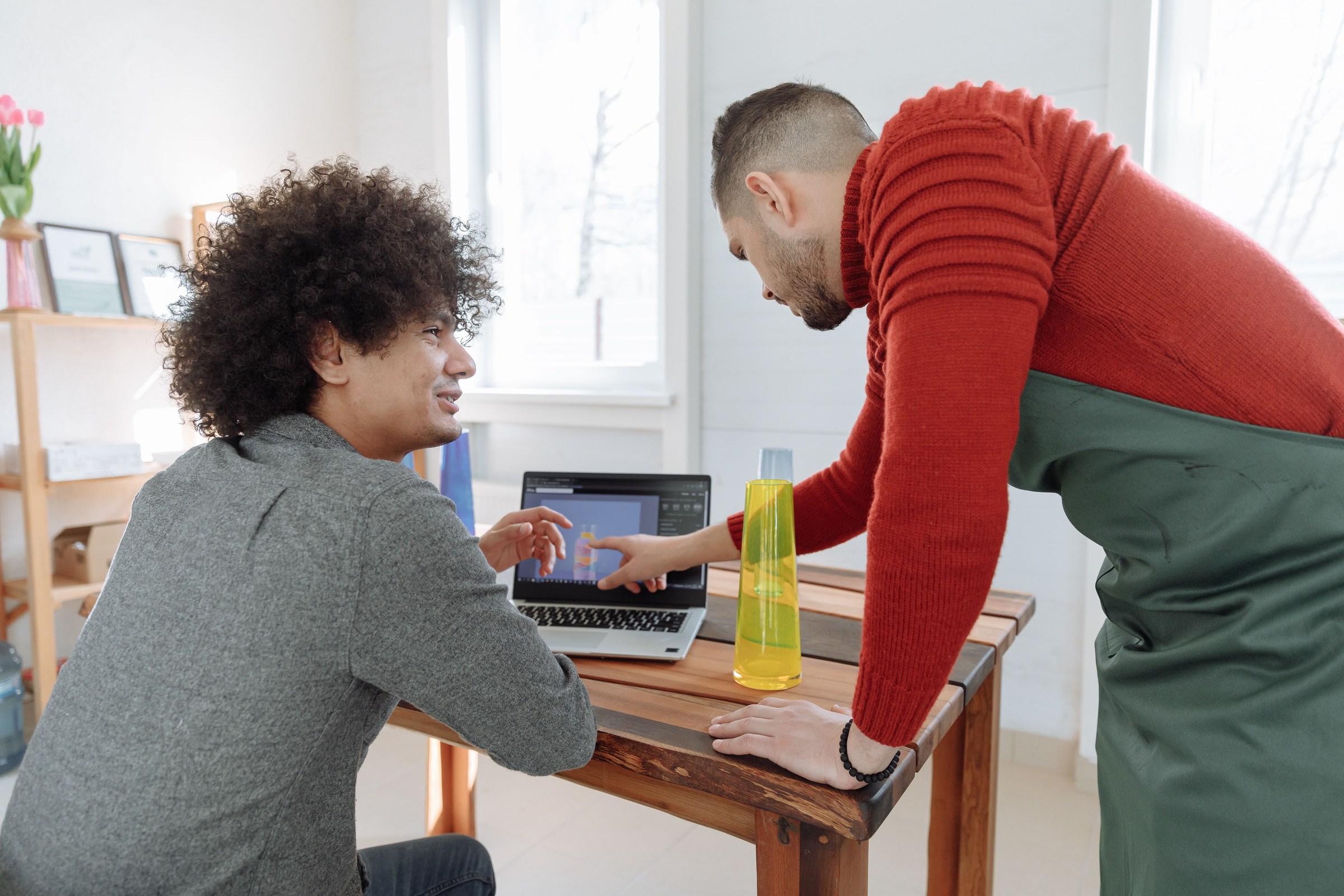 Shop owner in a green apron leaning over a desk with a younger marketer as they look at the first 30 days of work on a laptop