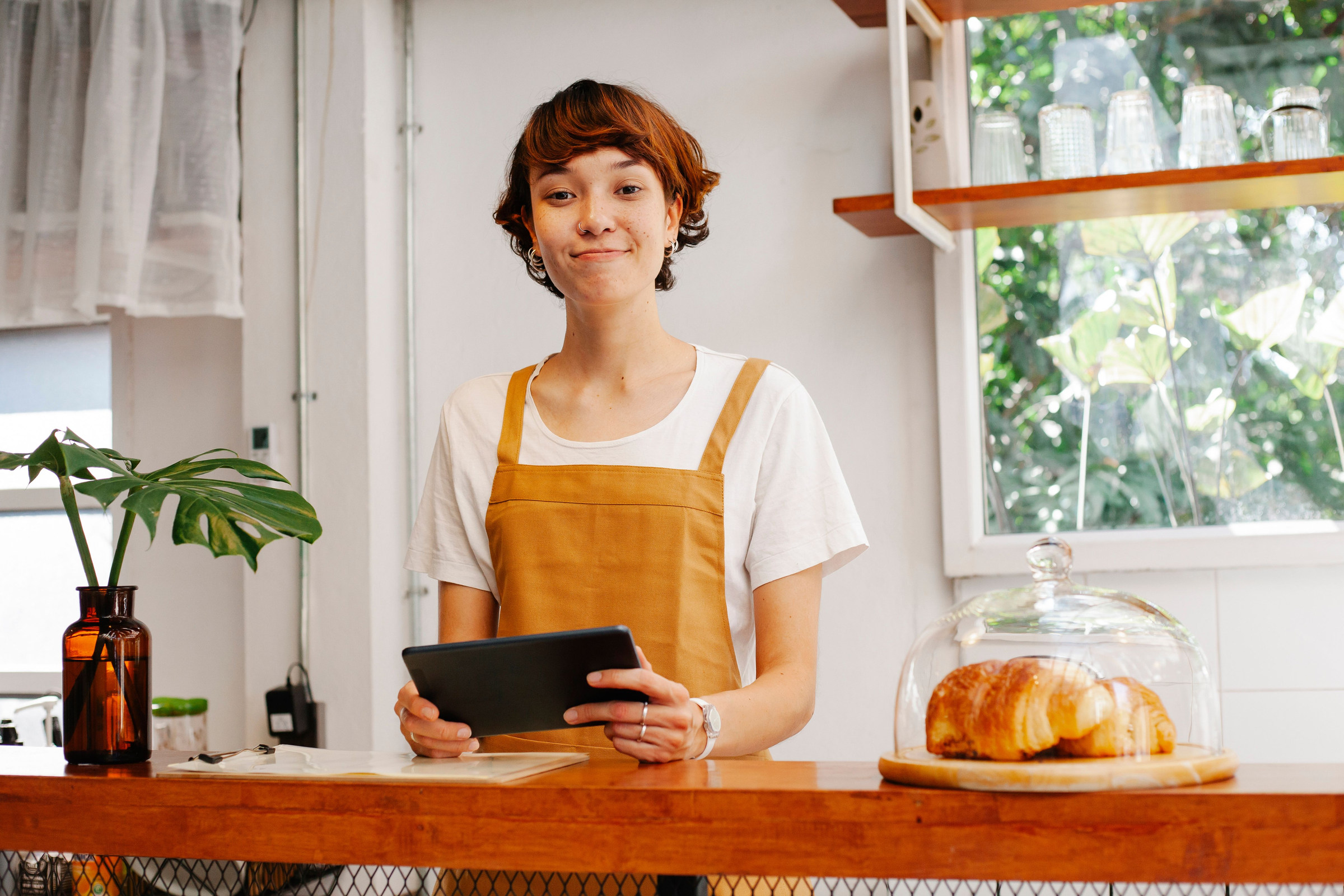 Smiling bakery employee in an apron holding a tablet at the counter beside fresh pastries, doing a quick monthly check of calls and leads