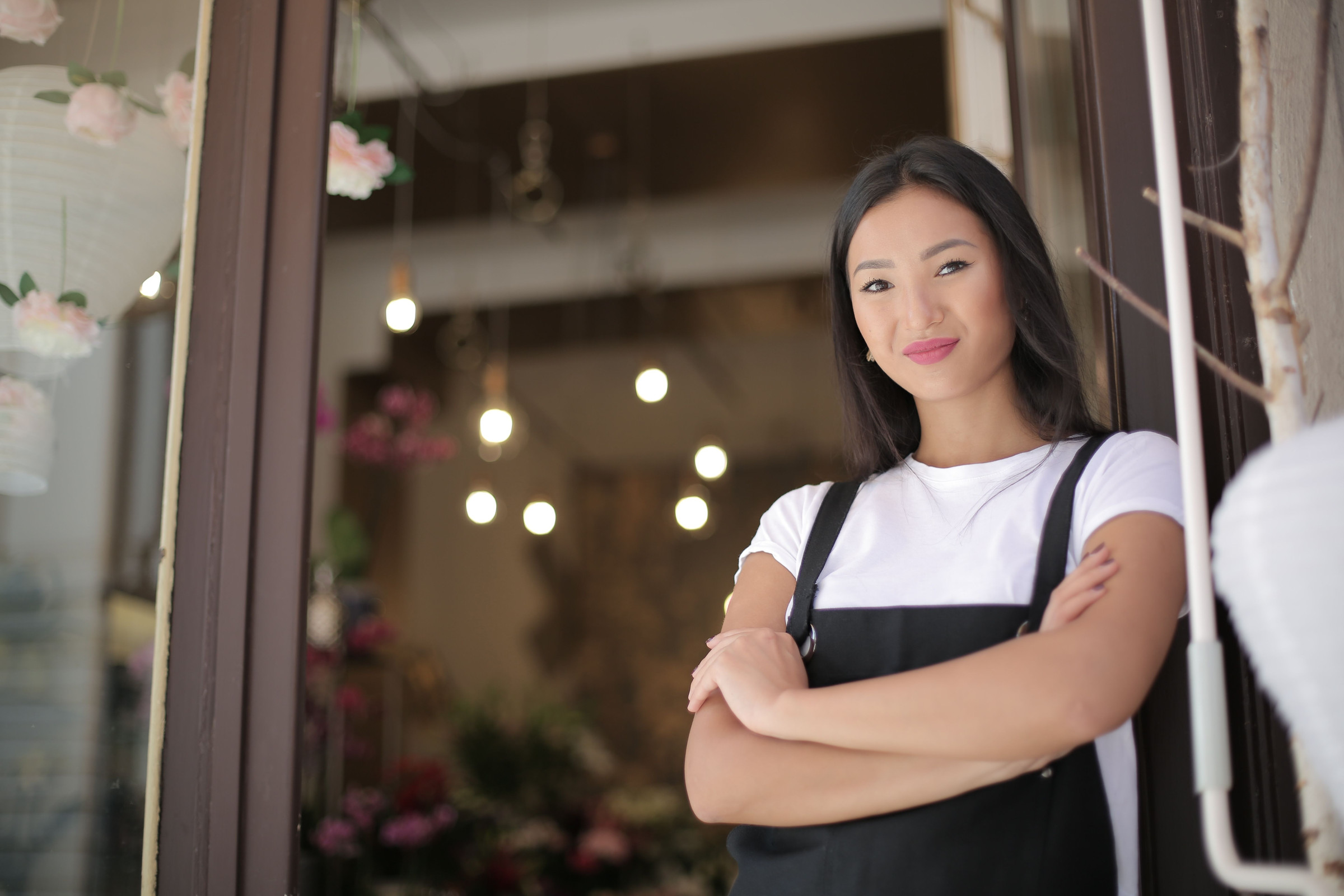 Smiling flower shop owner in a black apron standing with her arms crossed at the door of her shop, calm and confident