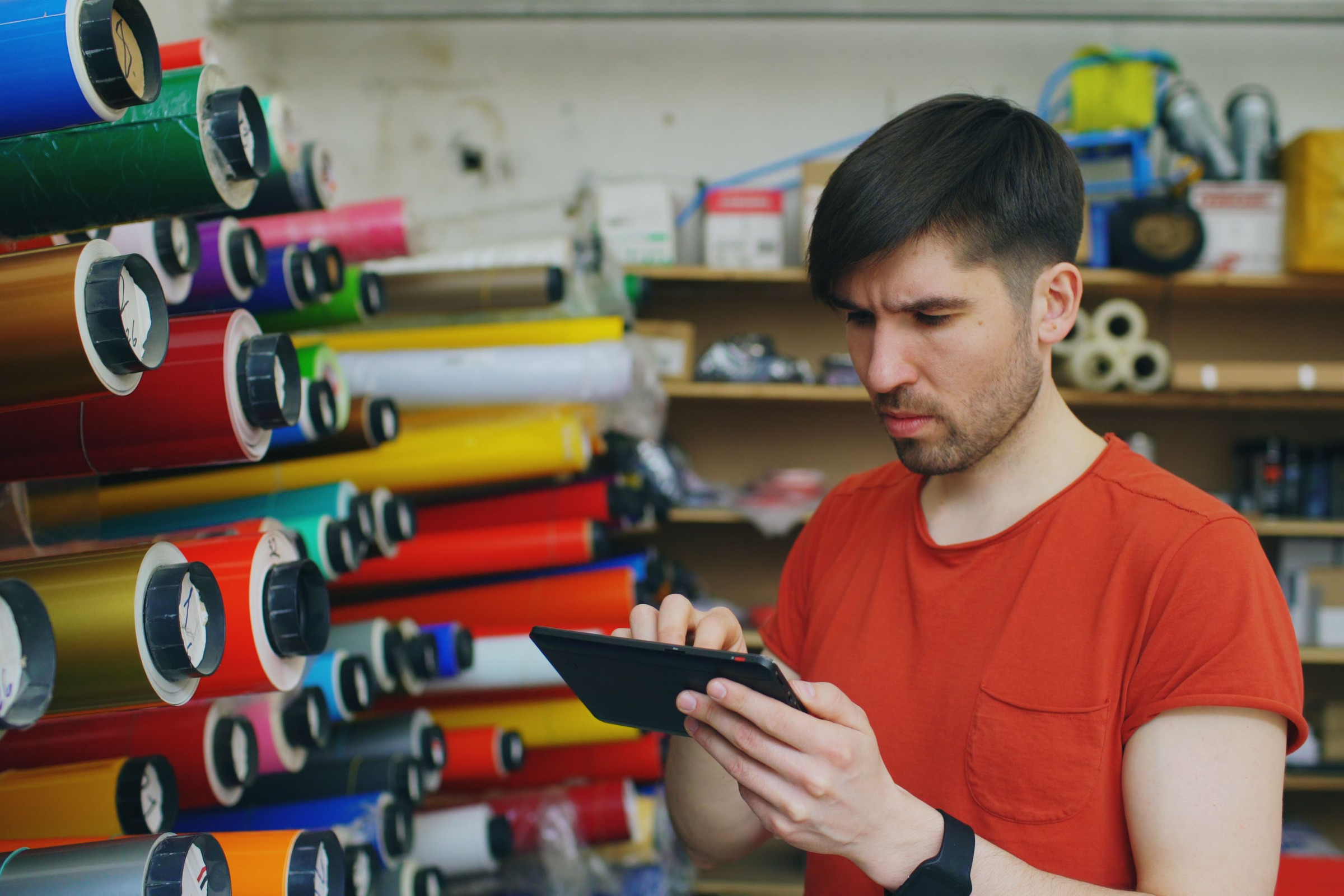 Print shop owner in a red shirt checking his Search Console numbers on a tablet in front of shelves of colorful vinyl rolls