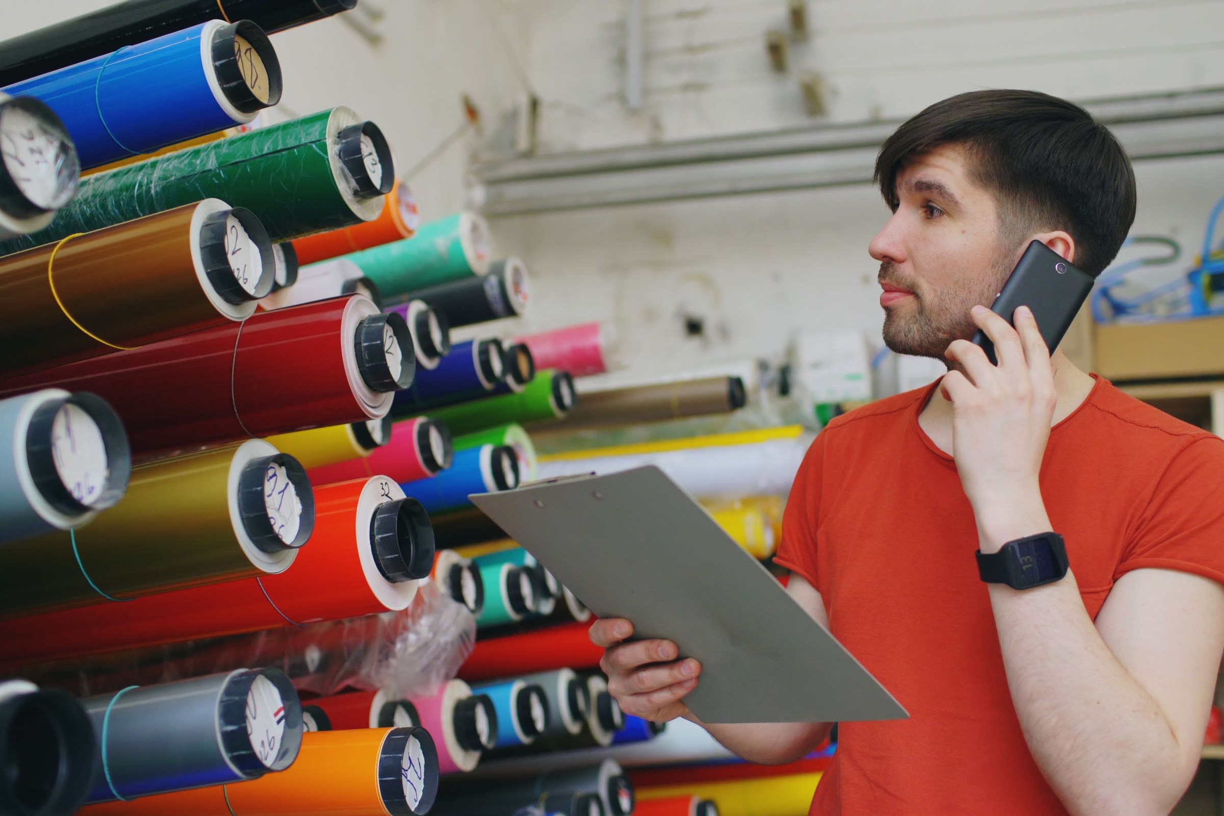 Print shop owner in a red t-shirt holding a clipboard and taking a customer call on his phone in front of shelves of colorful vinyl rolls