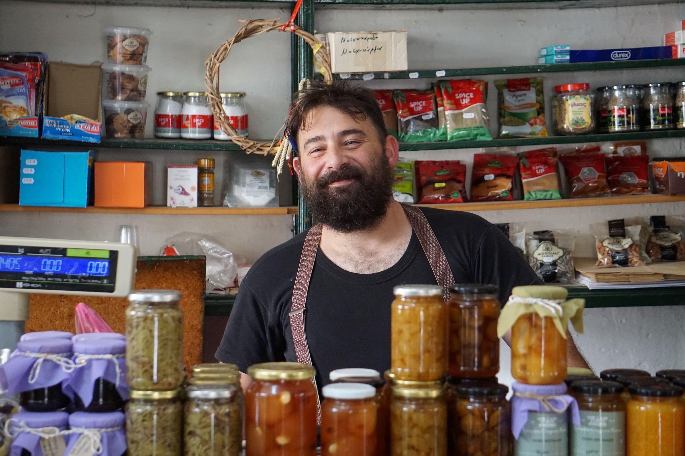 Bearded local shop owner in a black apron standing behind a counter of homemade preserves, representing a steady weekly or monthly listing and Knowledge Panel accuracy check