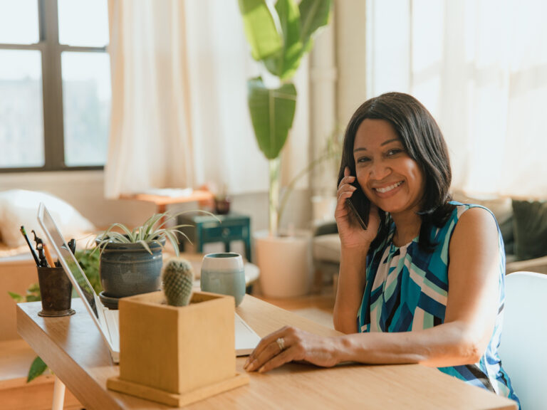 Smiling, polished small business owner in a teal patterned top taking a customer call at her desk in a bright, plant-filled office