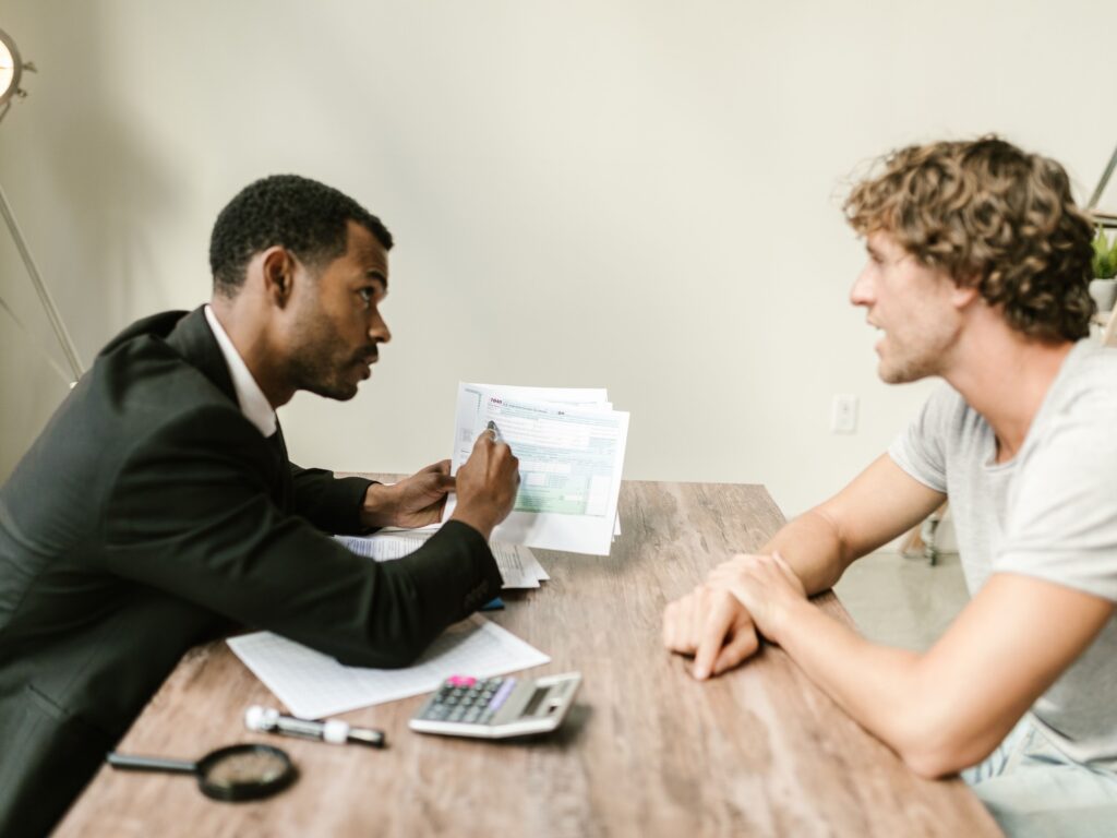 Local business owner in a t-shirt listening across a wooden desk as a marketing consultant in a suit walks him through printed paperwork, asking questions before he hires anyone