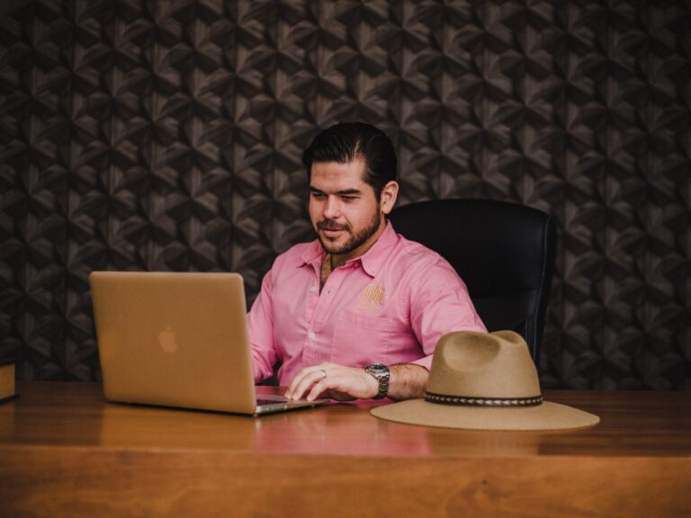 Small business owner in a pink shirt working at his wooden desk with a laptop and his hat beside him, checking how his website shows up in Google AI answers