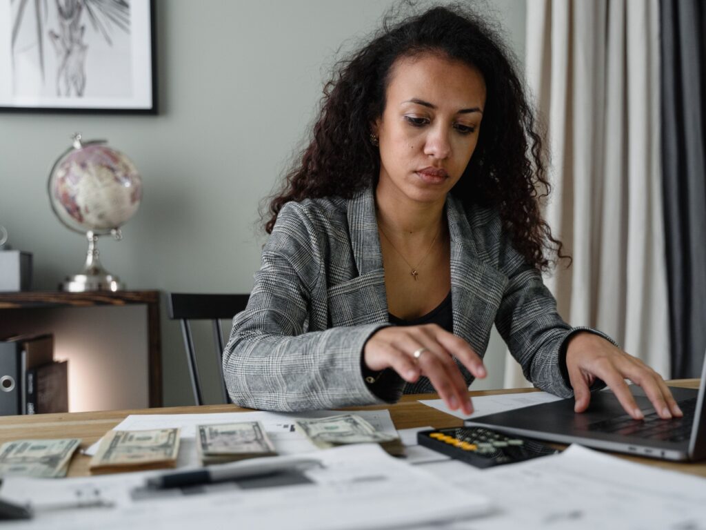 Local business owner in a gray plaid blazer at her desk with a laptop, calculator and receipts, reviewing what her Google ads cost and which leads turned into jobs