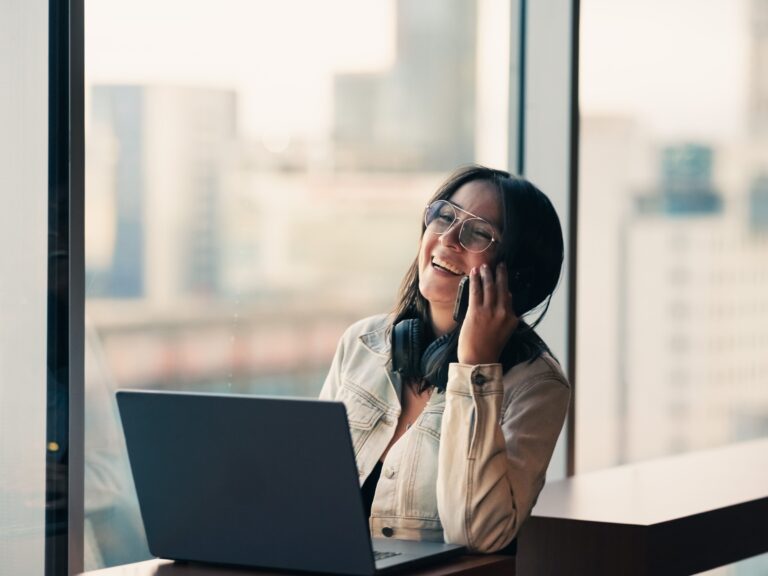 Smiling local business owner with glasses laughing on a phone call at a desk with a laptop, ready to check what Google’s AI Overview says about the business on mobile