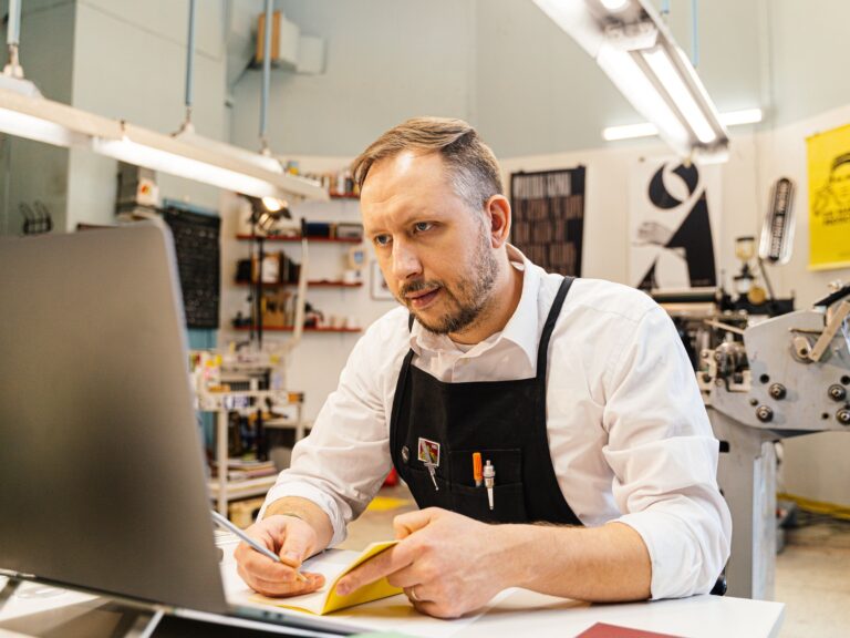 Local shop owner in a black apron at his workbench with a laptop and notepad, comparing his monthly Google ranking report with his own call and job numbers