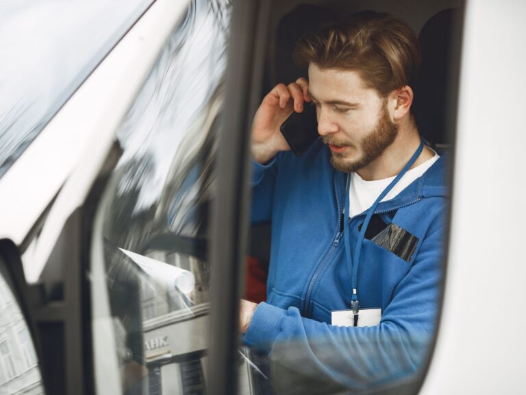 Tradesman in a blue work shirt sitting in the driver's seat of his work truck, taking a new customer call on his phone between jobs